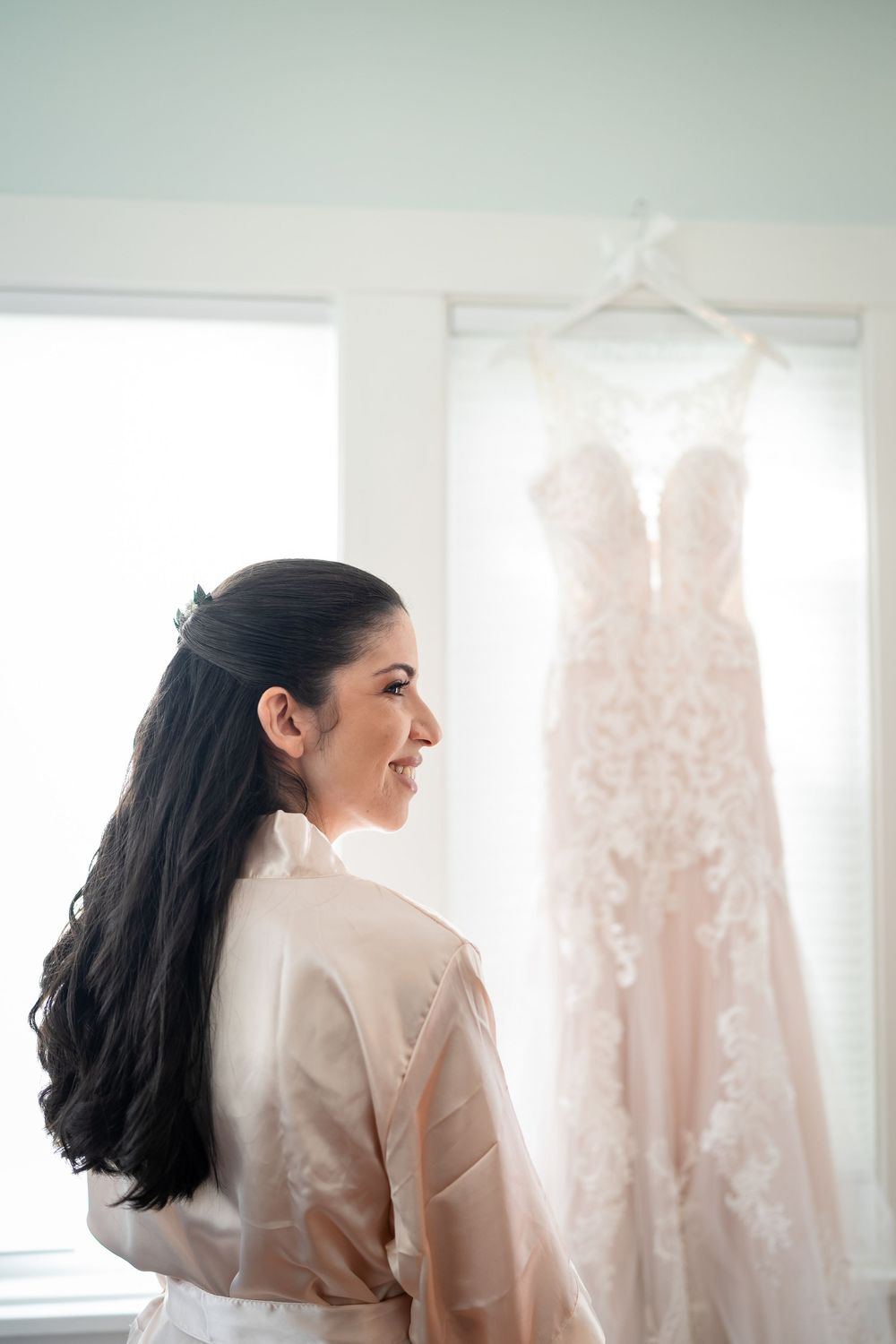A side view showing a blush silk robe against a backdrop of a hanging lace wedding dress by a window.