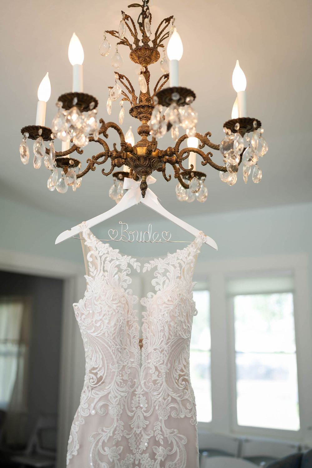 An elegant lace wedding dress hangs from a personalized hanger beneath an ornate crystal chandelier.