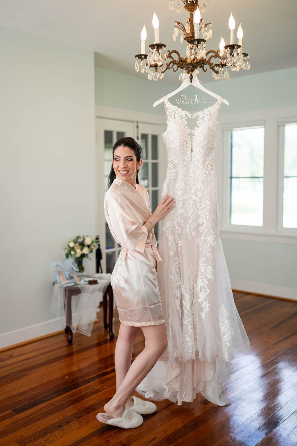 A beautiful lace wedding dress hangs from a crystal chandelier as someone in a silk robe stands beside it.