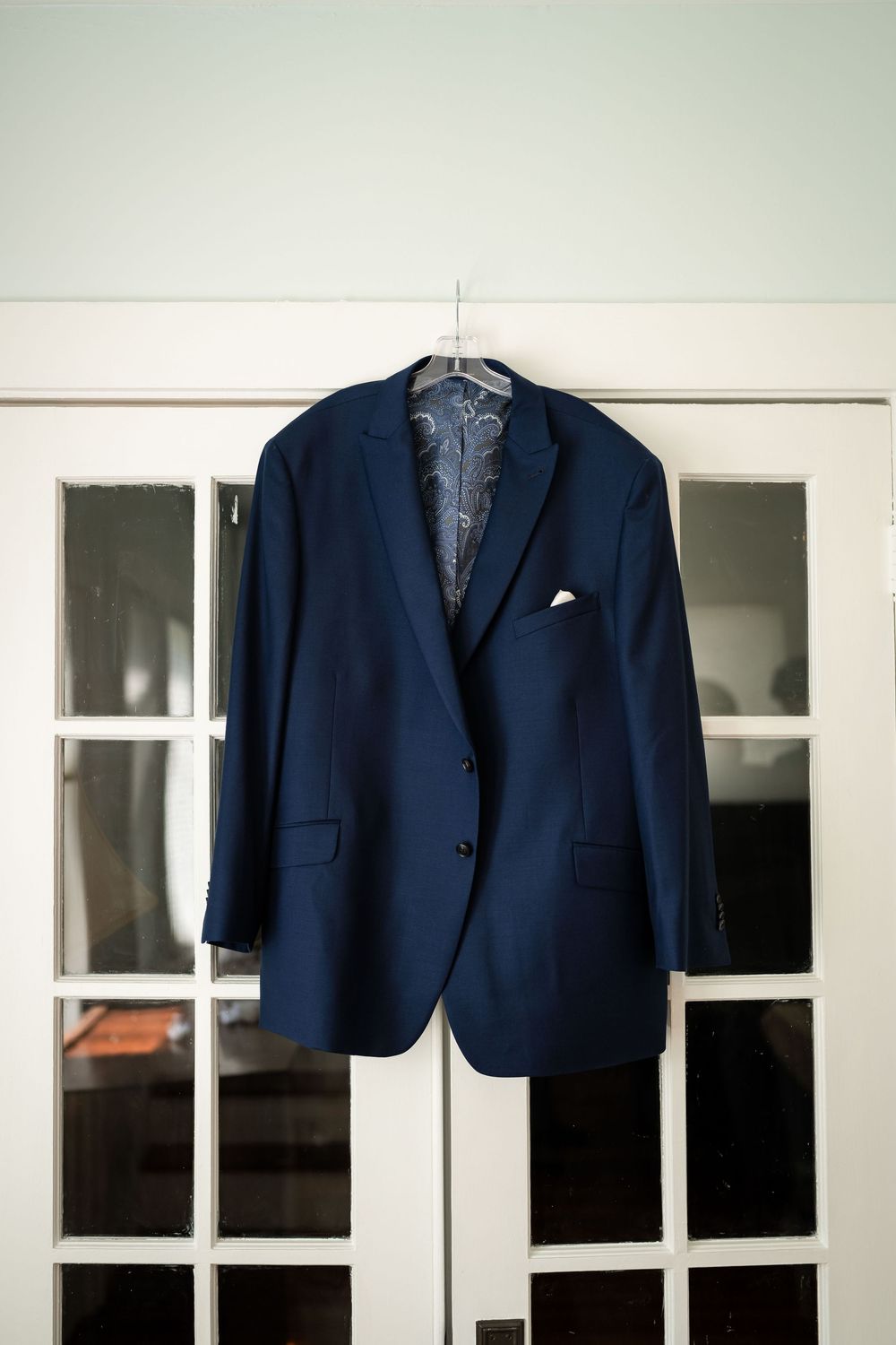 A classic navy blue men's suit jacket with a white pocket square hangs on display against French doors.
