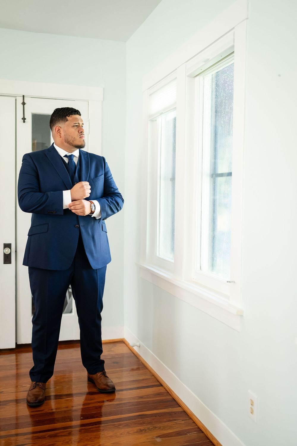 A groom in a navy blue suit adjusts his cuffs while standing in a bright room.