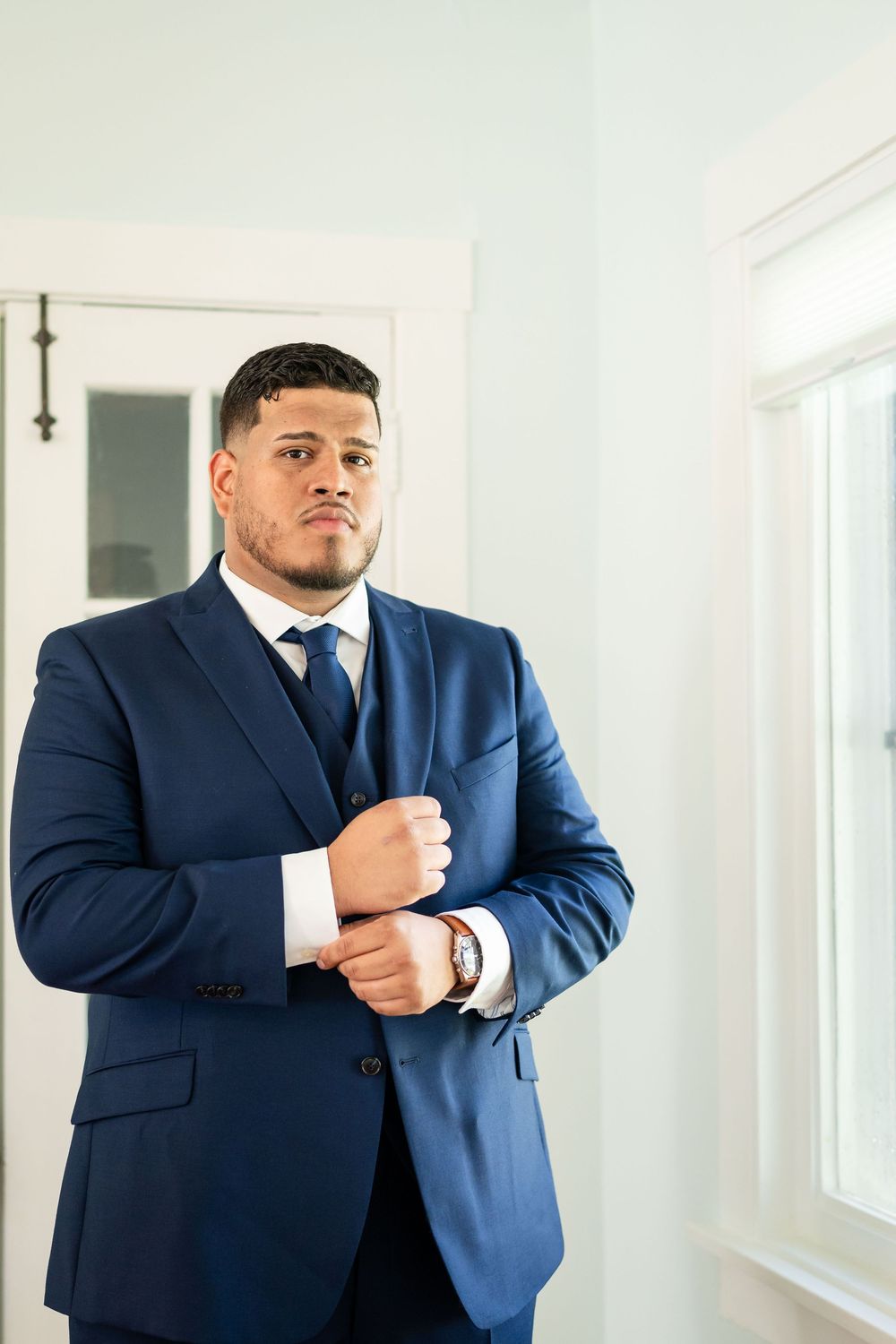 A person in a navy blue suit adjusts their cuff while standing in a bright white doorway.
