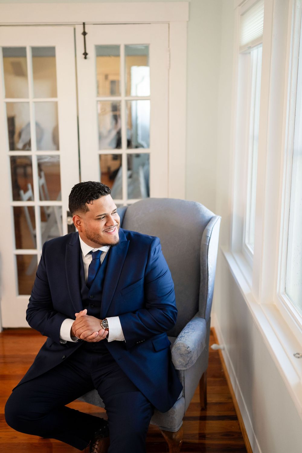 A person wearing a navy blue suit sits in an elegant gray armchair by a bright window in a light-filled room.