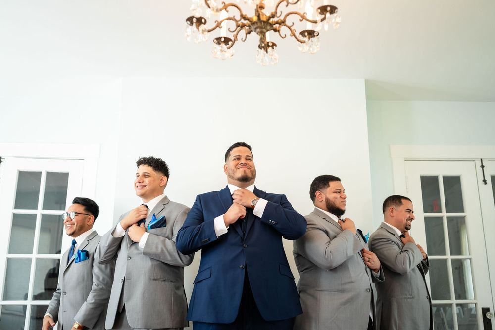 A groom in navy blue suit stands with groomsmen in grey suits adjusting their ties while getting ready.