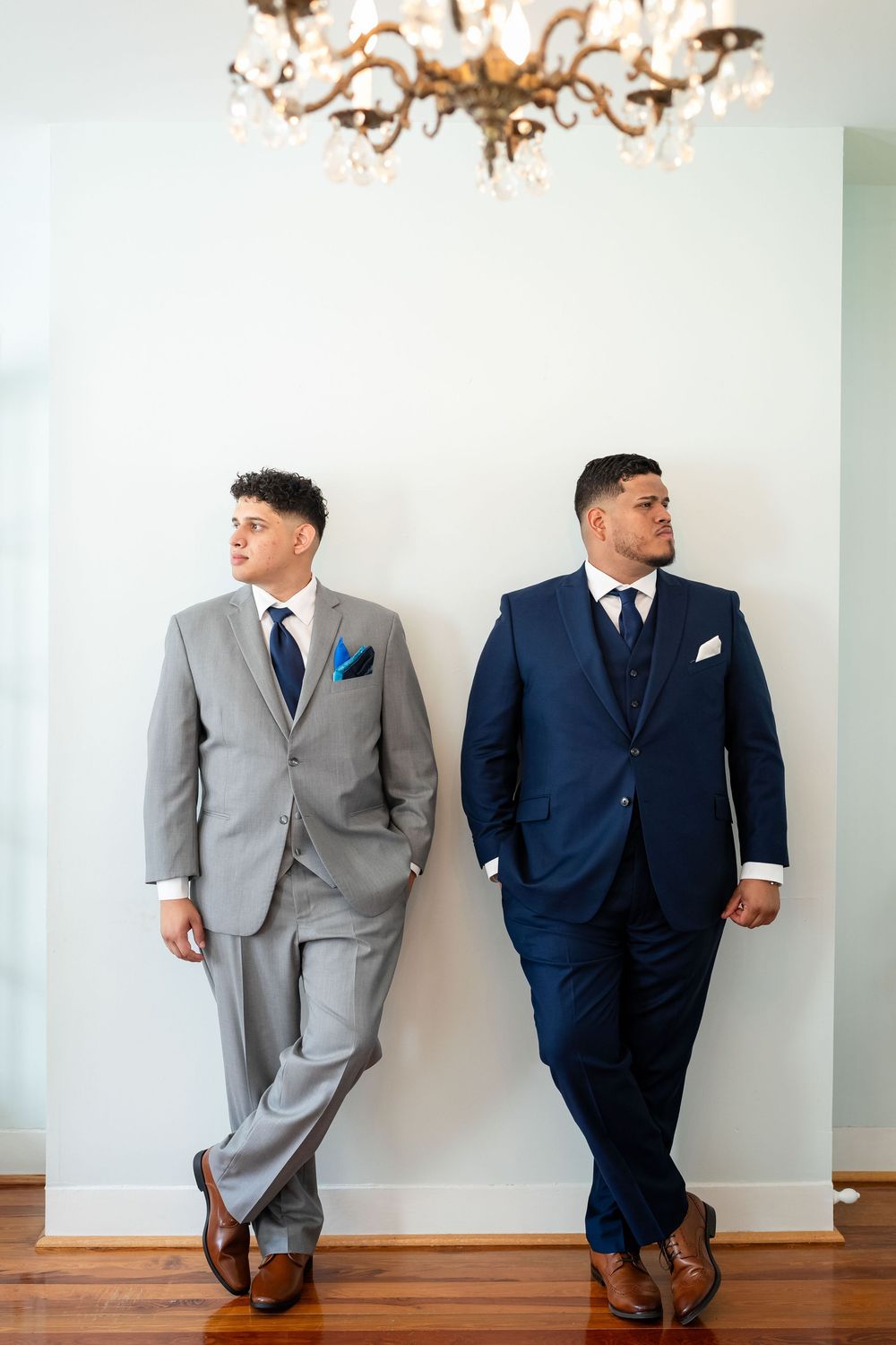 Two people in formal suits - one in gray and one in navy - lean against a white wall under an elegant chandelier.