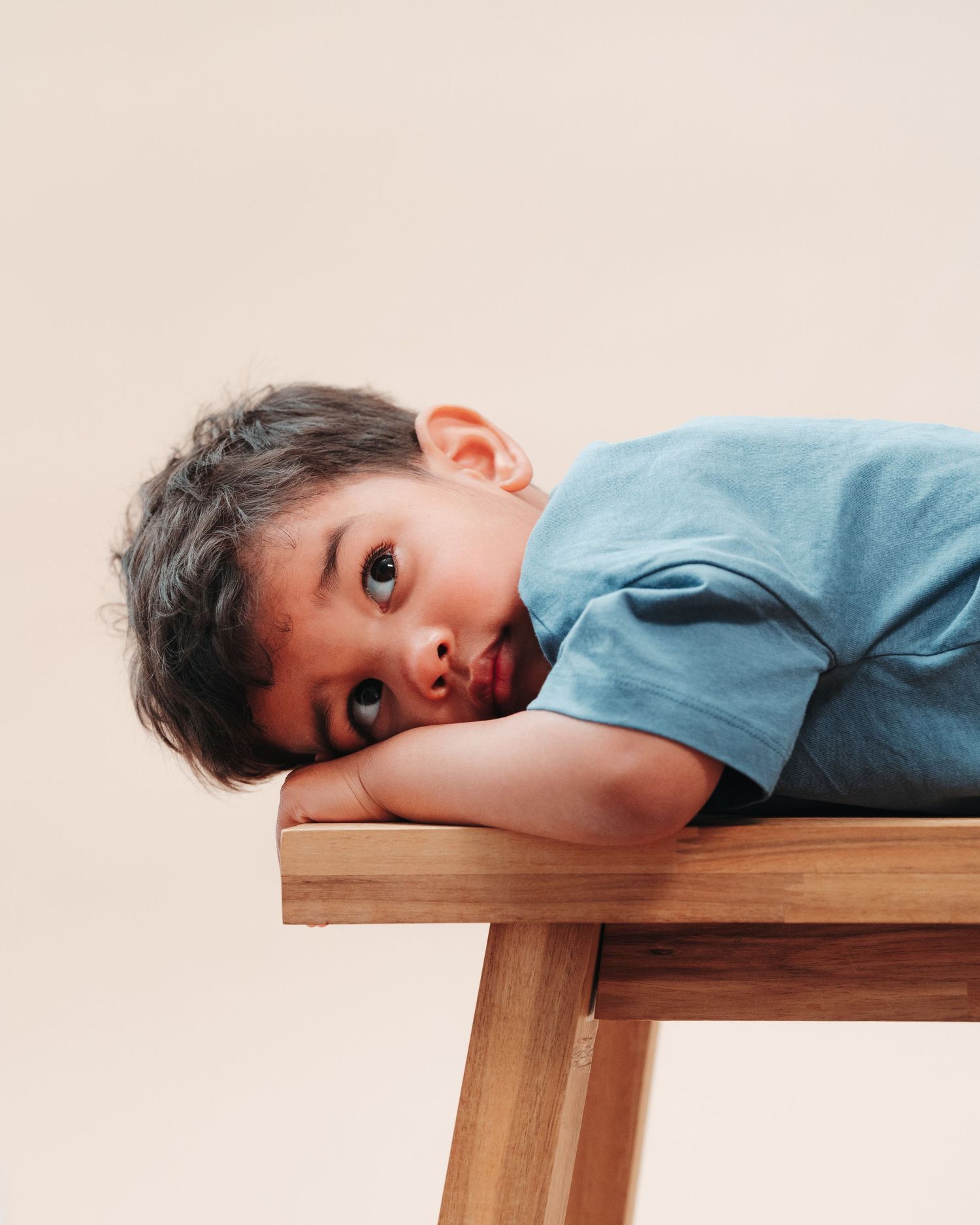 Child resting head on wooden surface with curious expression against pink backdrop.