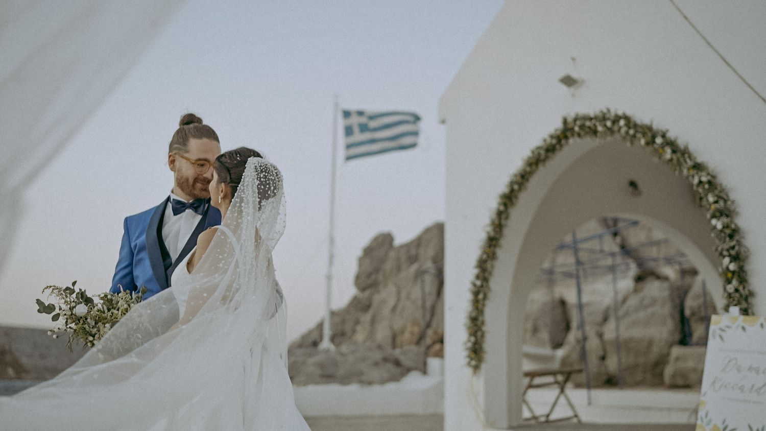 A bride with flowing veil stands under a white archway in Greece with mountains and Greek flag visible in background.