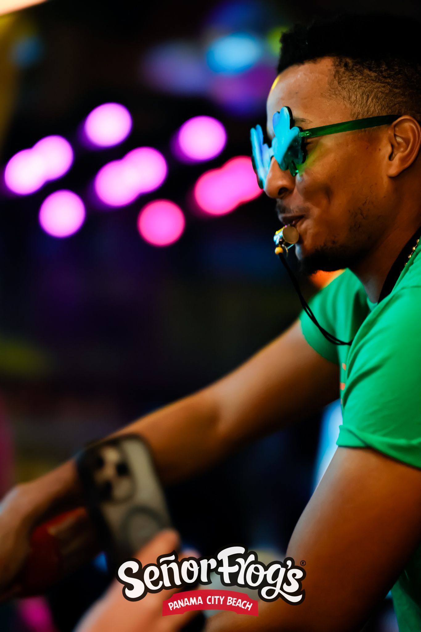 DJ wearing bright green shirt and neon sunglasses performing at club.