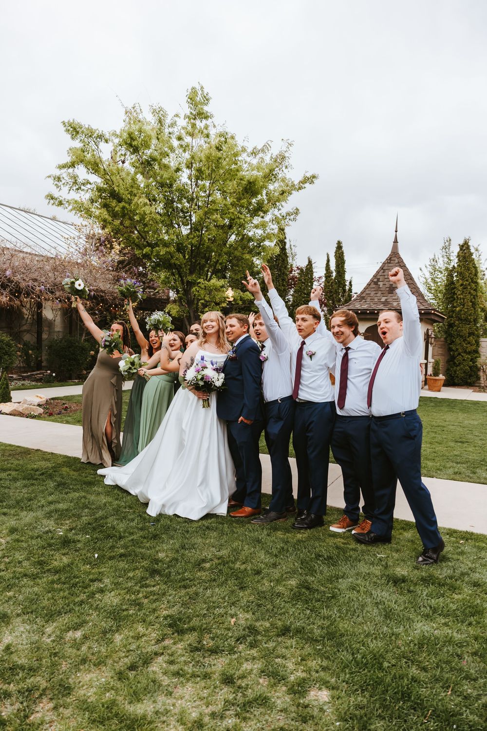 Wedding party posing joyfully on a lawn with a chapel in the background.