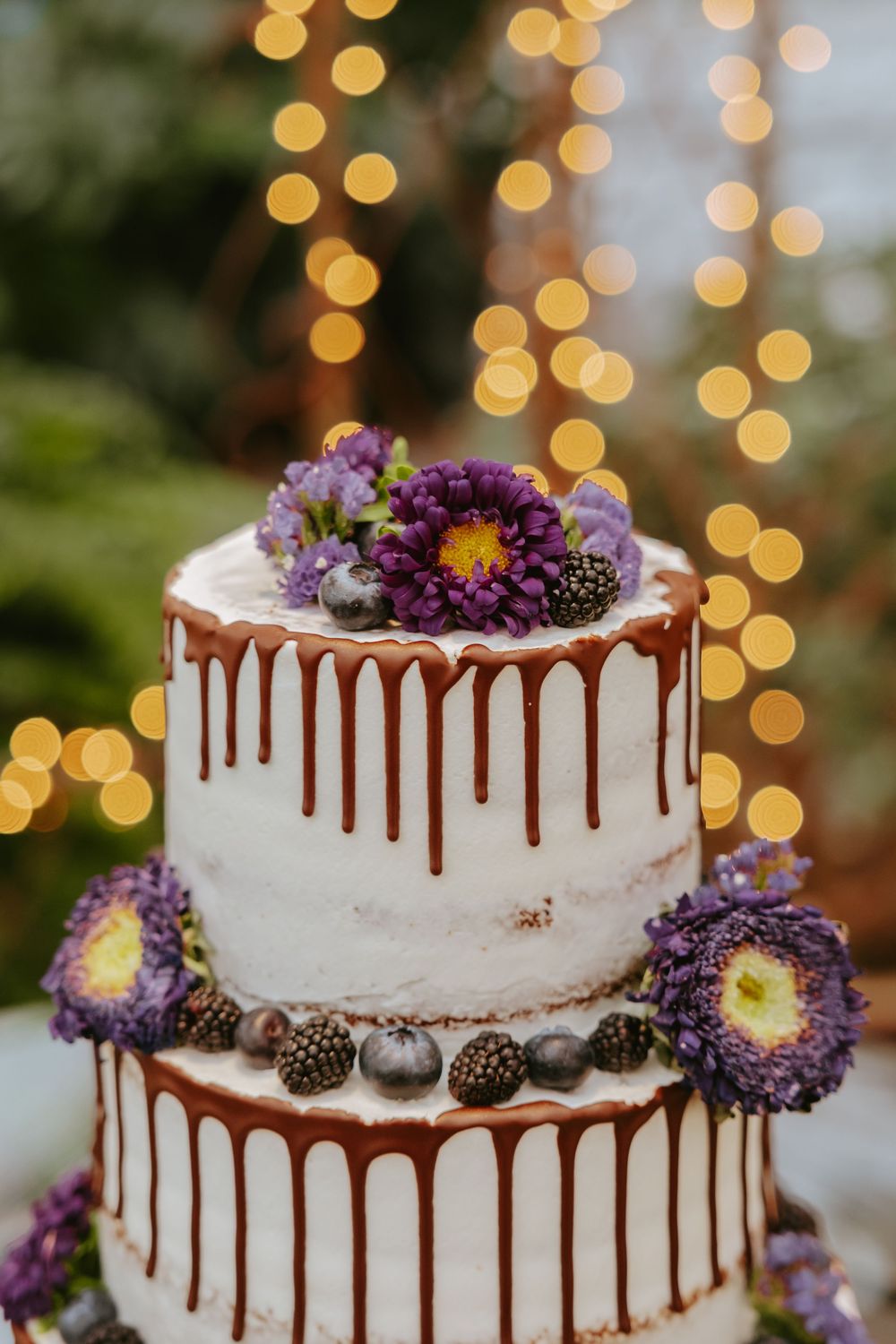 Two-tier white wedding cake with chocolate drip and purple flowers decorated against a soft bokeh background.