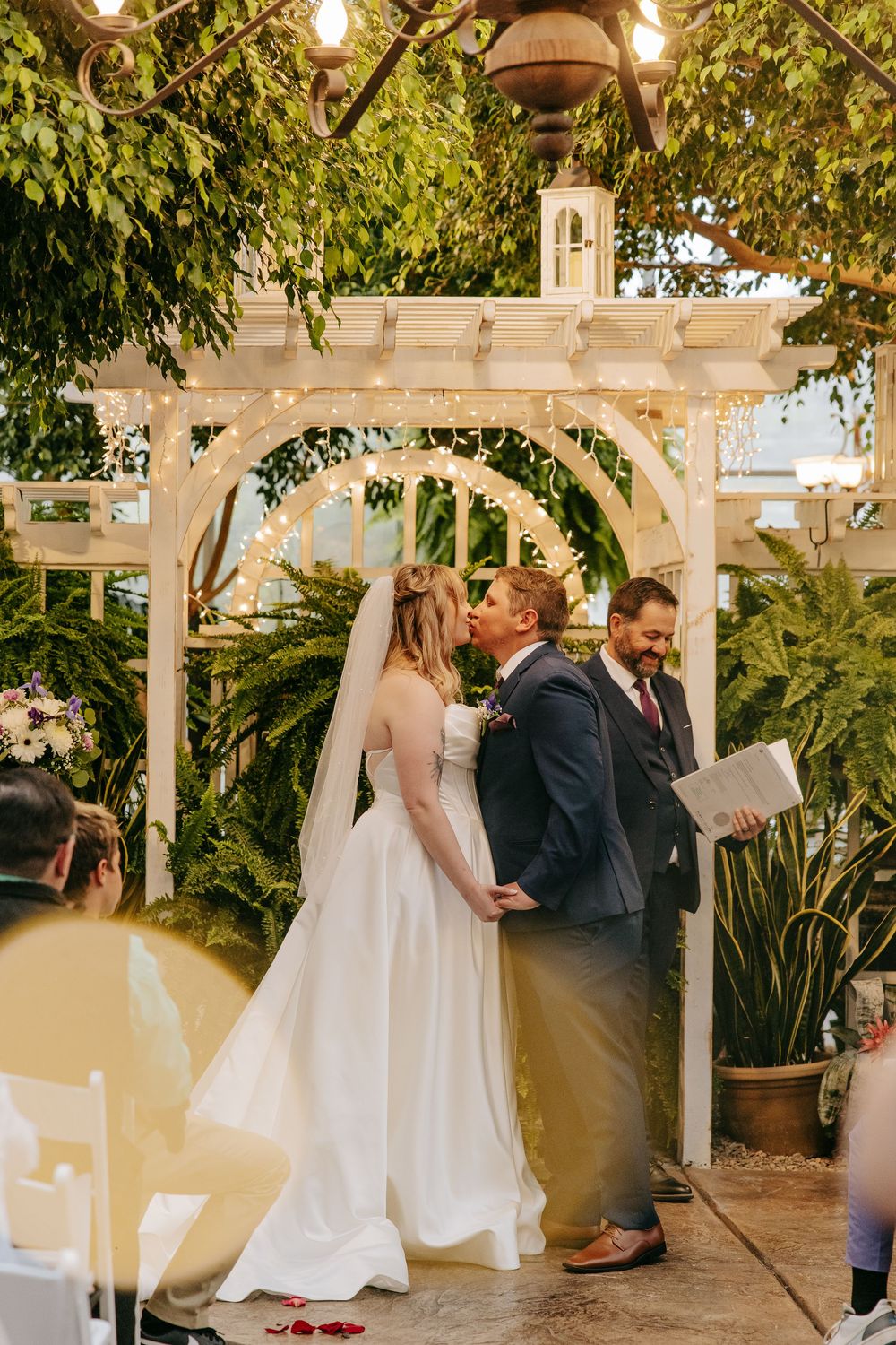Couple exchanges vows under a white pergola decorated with string lights and lush greenery.
