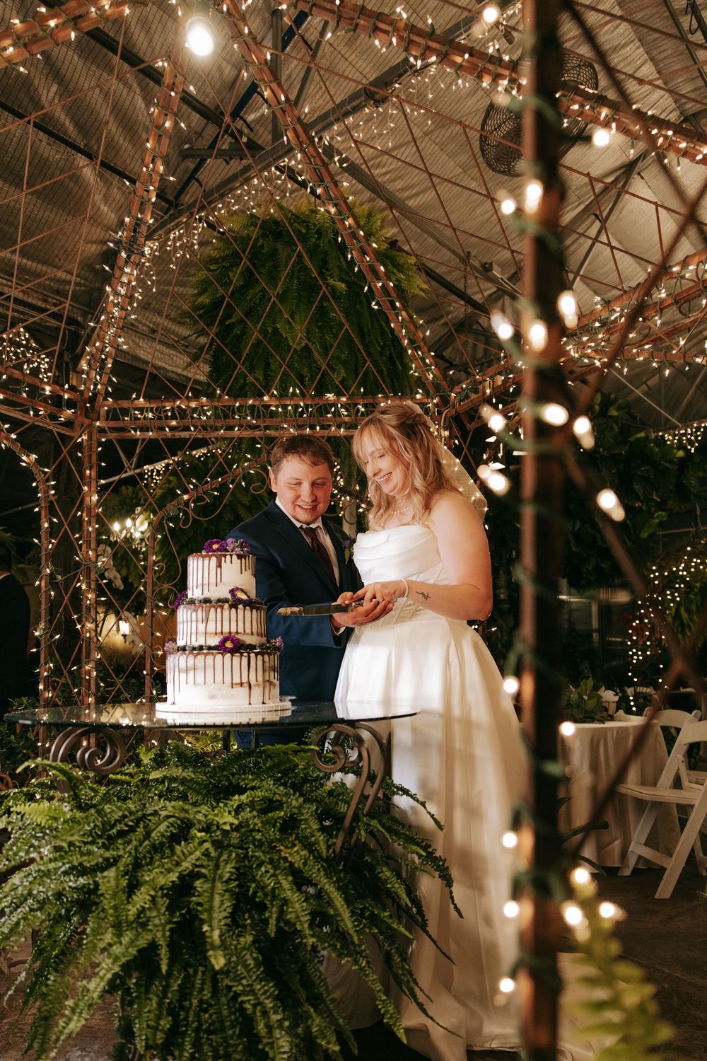A wedding couple prepares to cut their drip-style tiered cake beneath an enchanting canopy of twinkling lights and greenery.