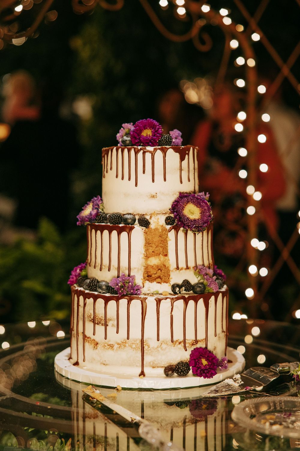 Elegant three-tier white wedding cake decorated with chocolate drip and purple flowers against twinkling lights.