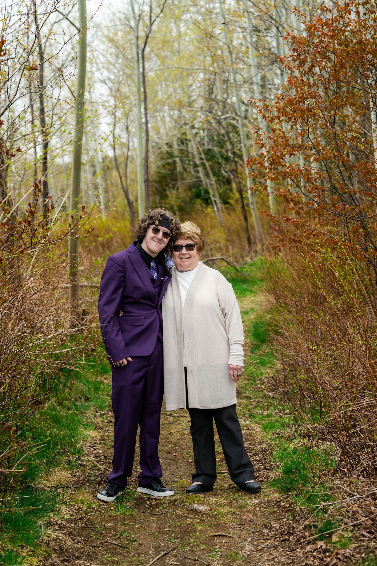 Two people in contrasting outfits stand together on a woodland trail during autumn with bare trees in background.