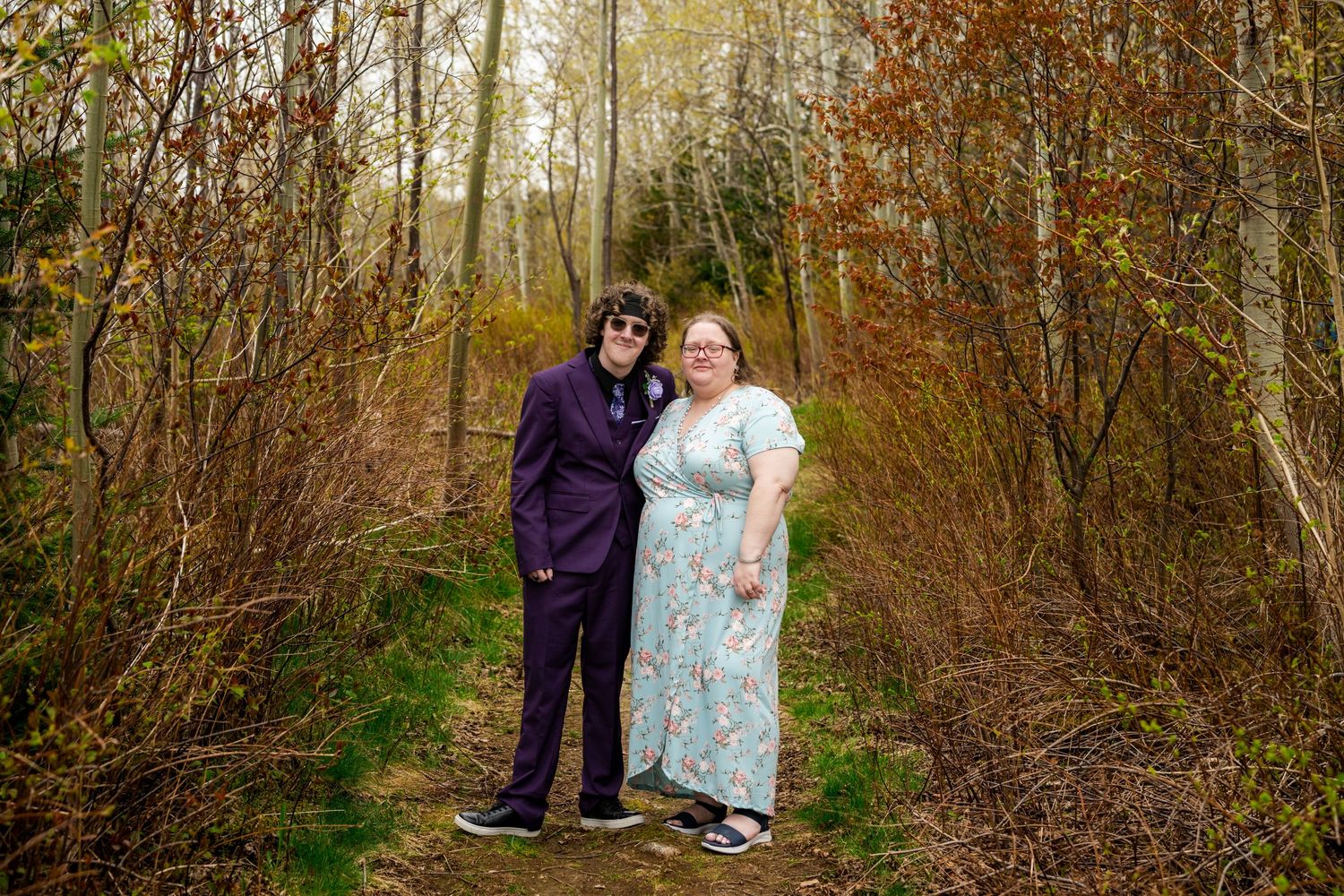 Two people in contrasting formal attire stand together on a forest path during autumn.