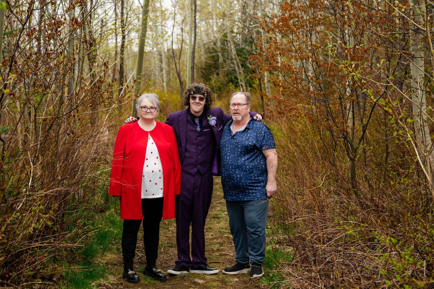 Three people stand together outdoors on a nature path surrounded by autumn trees and foliage in the background.
