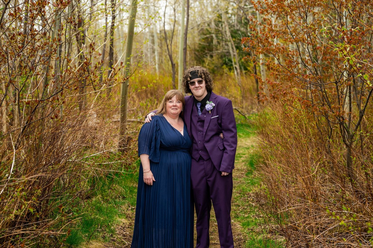 A couple in formal wear standing on a forest path with autumn foliage and golden light in the background.