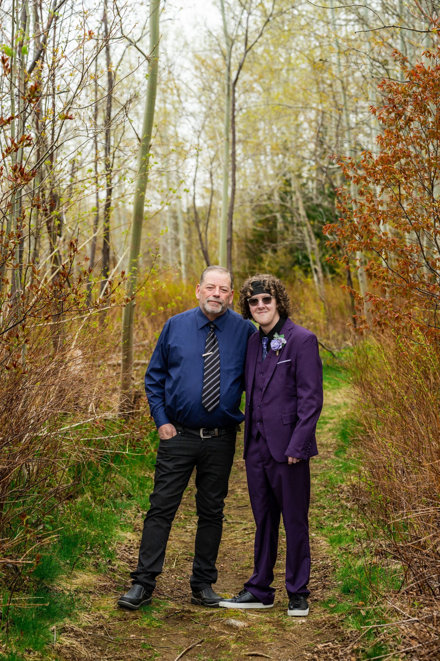 Two people in autumn attire stand on a woodland trail surrounded by brush and trees in golden fall colors.