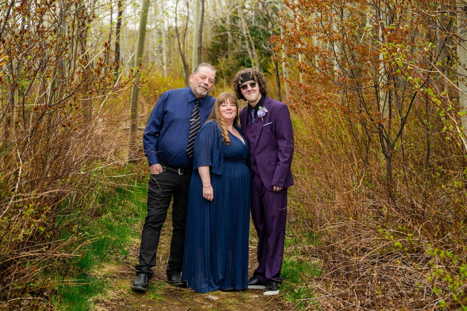 Three people pose together on a woodland path surrounded by autumn foliage and bare branches.