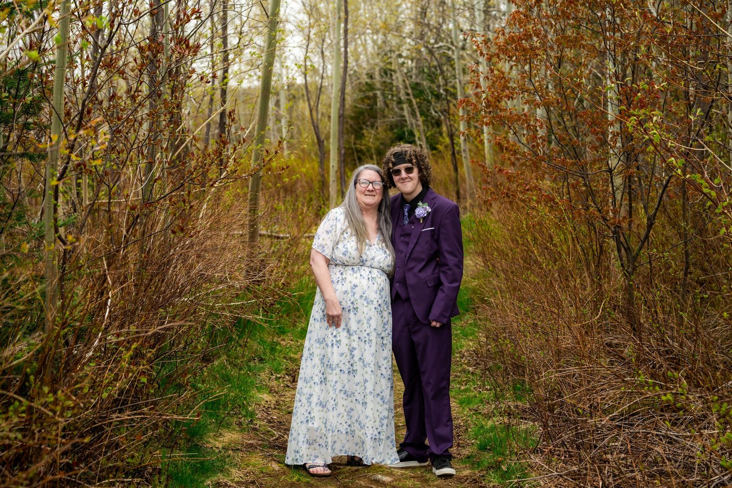 Two people in formal attire stand together on a wooded path surrounded by fall foliage.