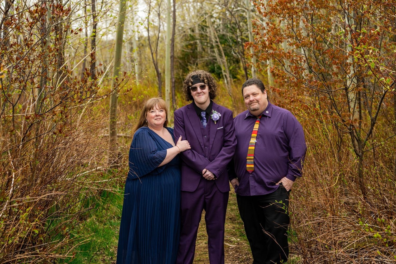 Group wearing coordinating blue and purple outfits poses together in front of autumn trees in natural setting.