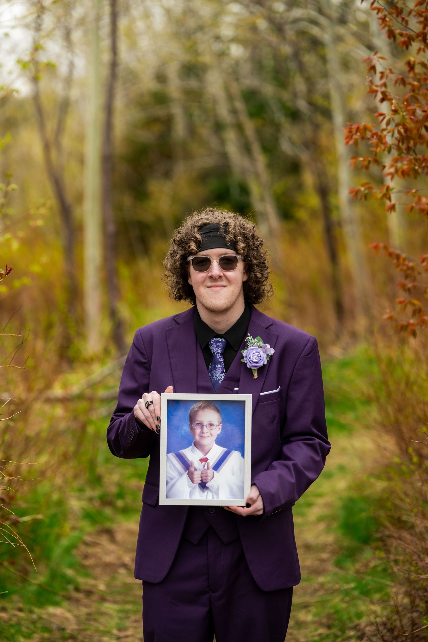 A person in a purple suit and sunglasses stands outdoors while holding a framed photograph amid fall foliage.