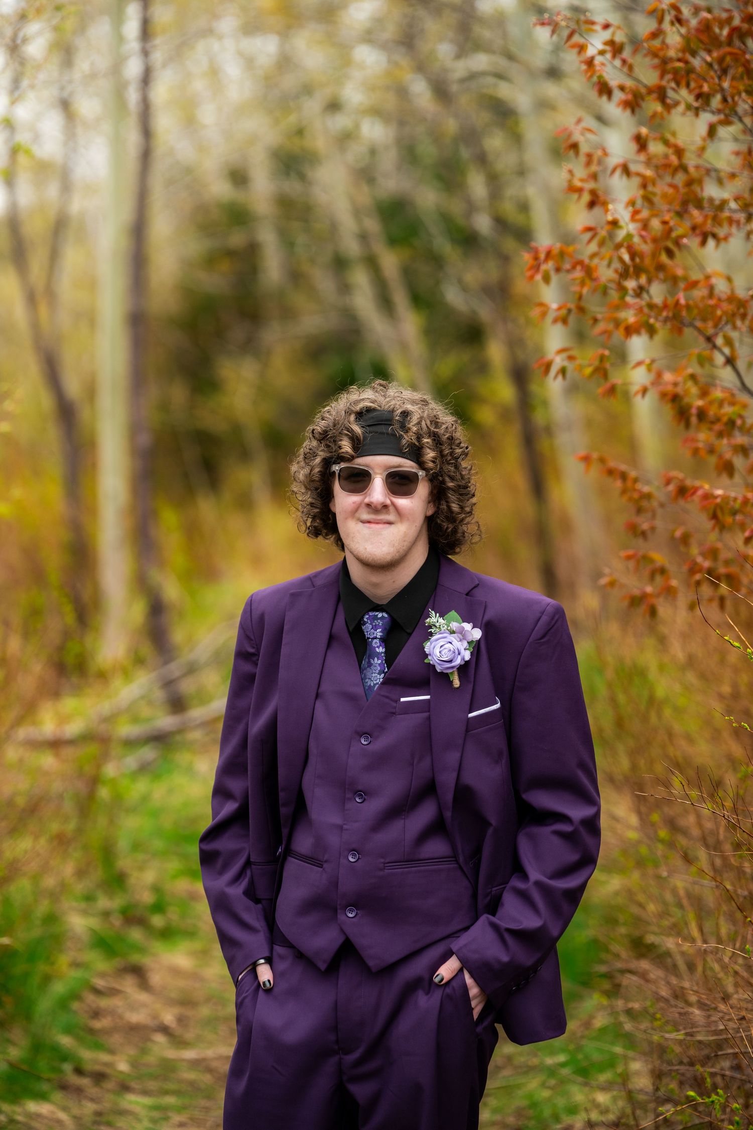 A person in a purple suit stands confidently on a wooded trail with autumn foliage in the background.
