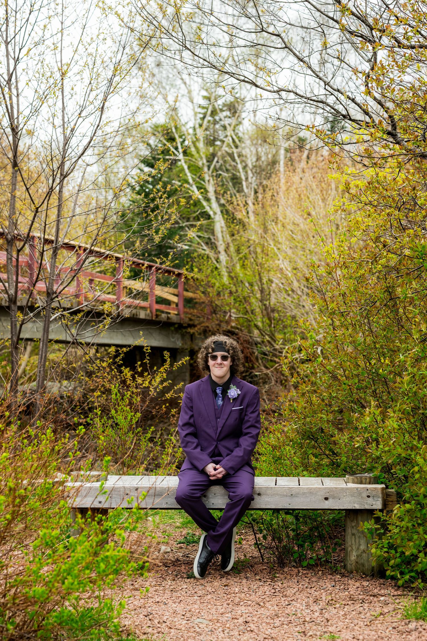 A wooden footbridge spans over a trail surrounded by autumn trees and bushes with yellow foliage in a park setting.