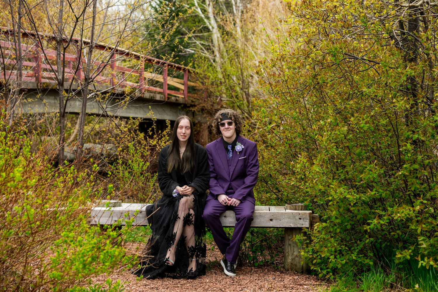 Two people sit together on a bench beneath a wooden footbridge surrounded by autumn foliage in a park setting.