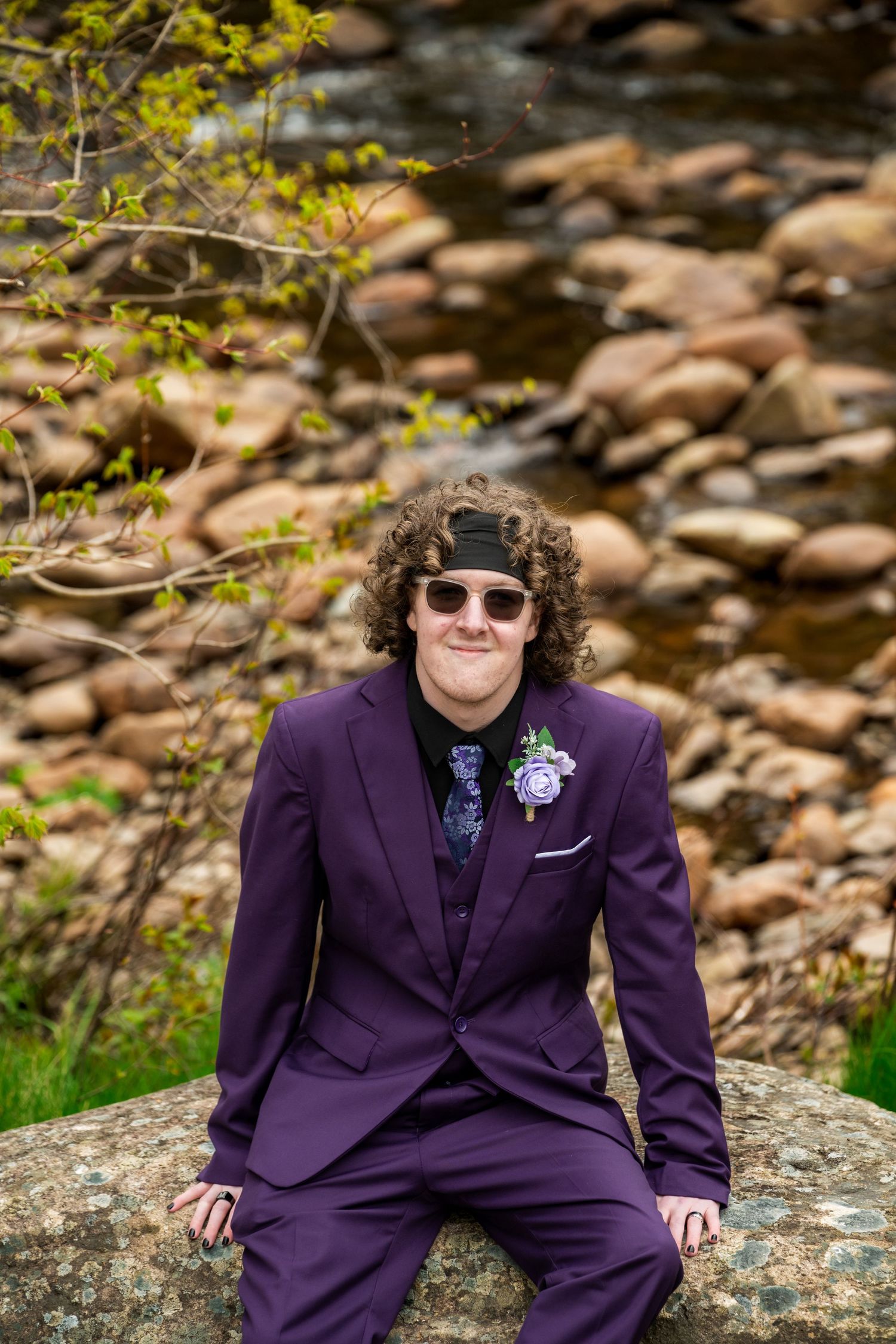 A well-dressed person in a purple suit sits on a rocky outcrop near a stream with sunglasses and a boutonniere.