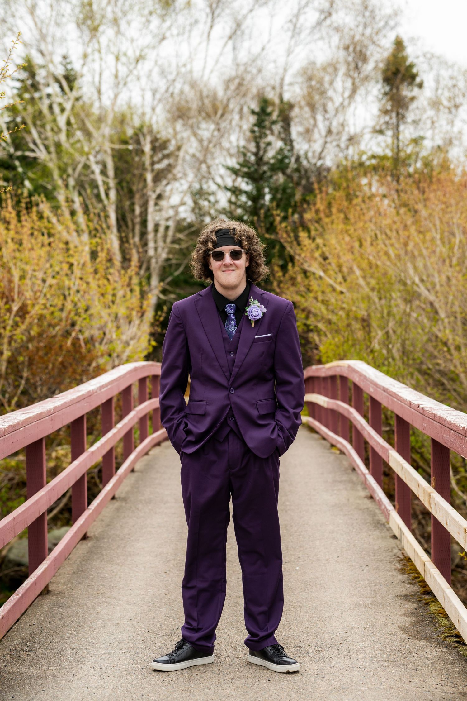 A person in a purple suit and sunglasses stands confidently on a wooden bridge surrounded by autumn foliage.