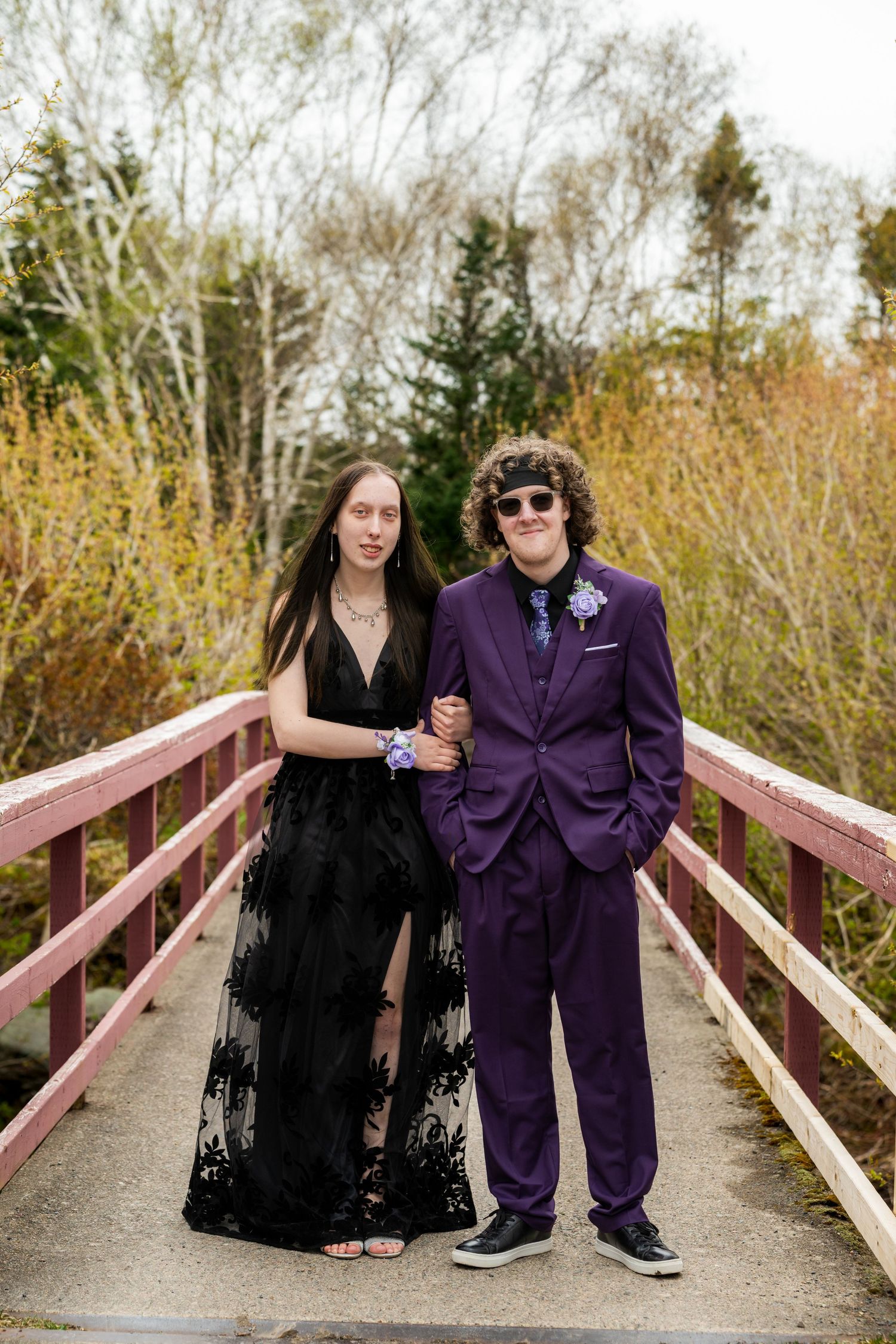 A couple in formal attire poses on a wooden bridge, with trees and autumn foliage in the background.