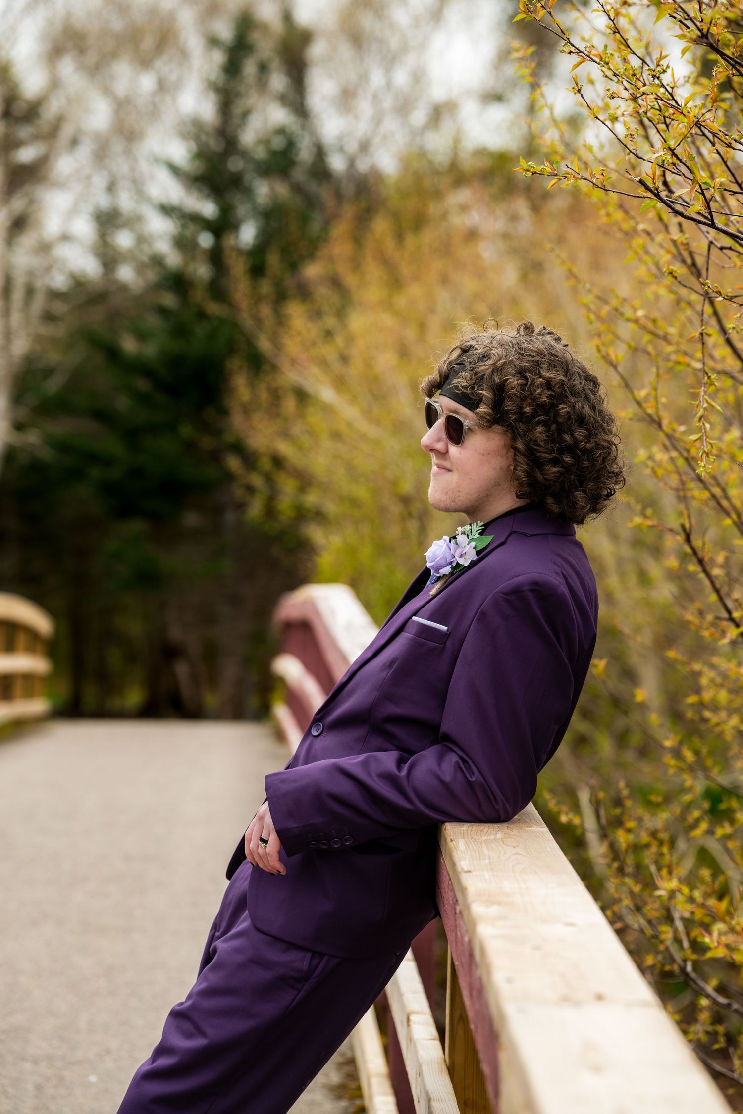 A person in a purple suit and sunglasses leans against a wooden bridge railing in an autumn outdoor setting.