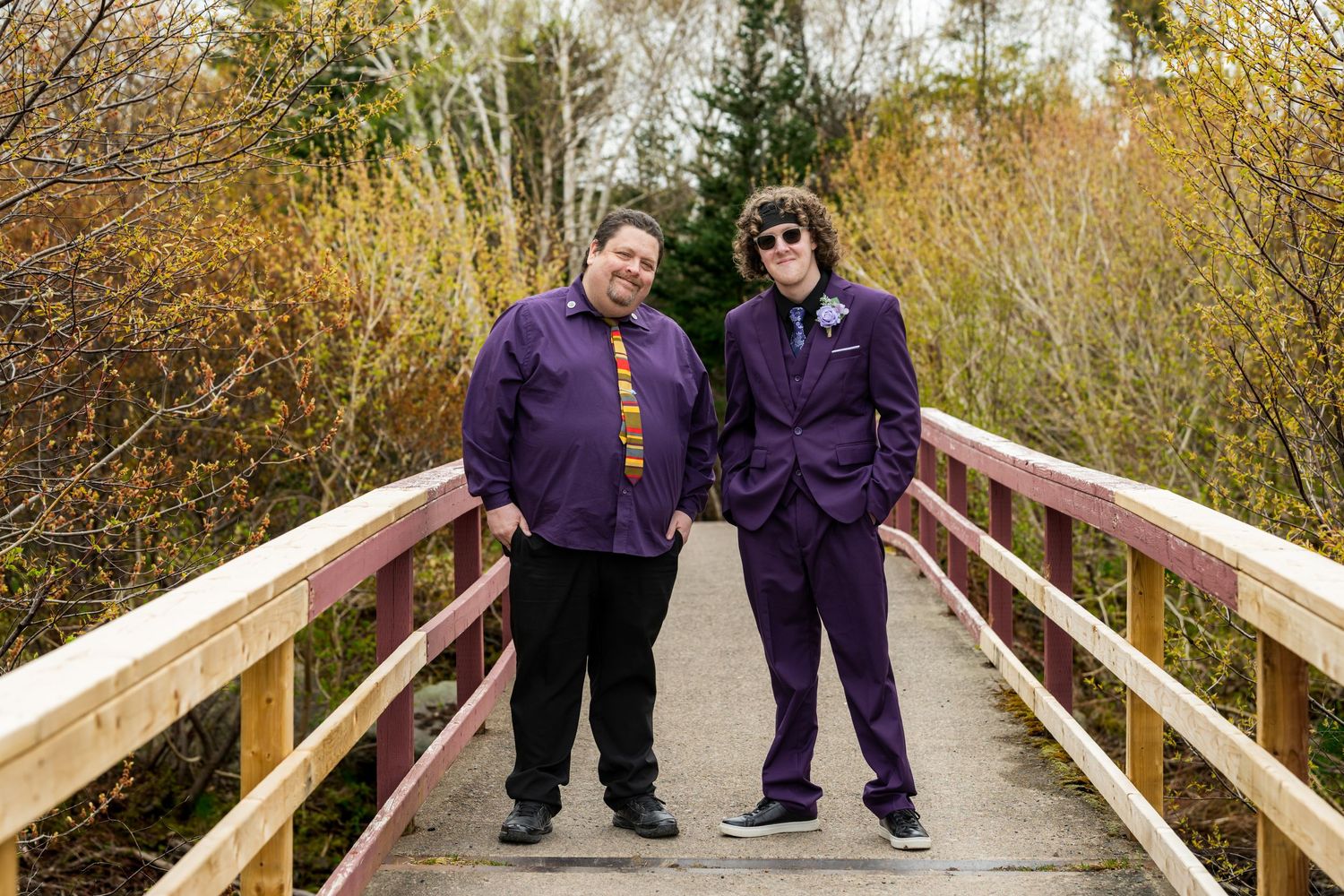 Two people wearing matching purple jackets stand together on a wooden footbridge surrounded by autumn foliage.