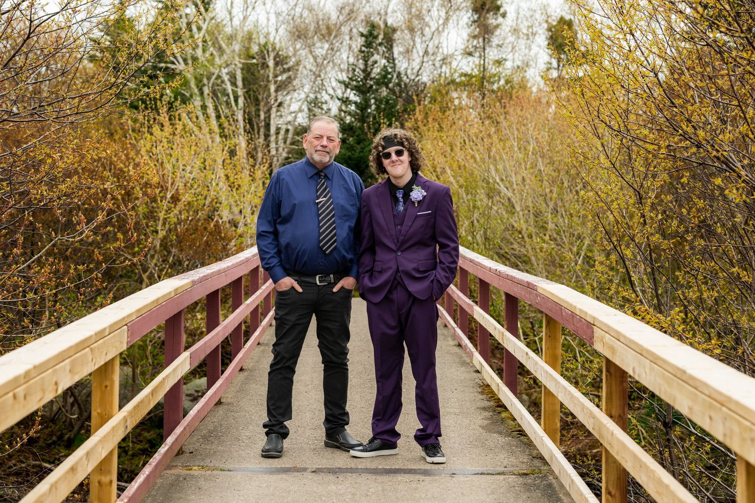 Two people in casual jackets stand together on a wooden footbridge surrounded by autumn foliage.