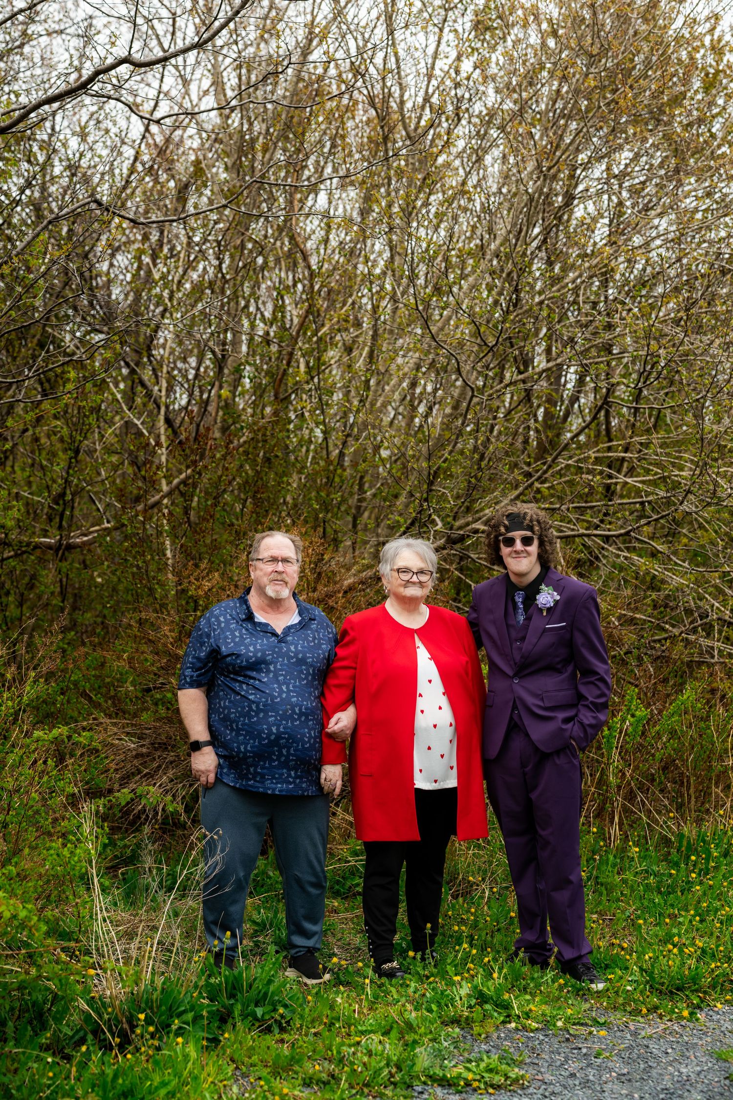 Three people stand together on a gravel path with brushy trees and greenery in the background.