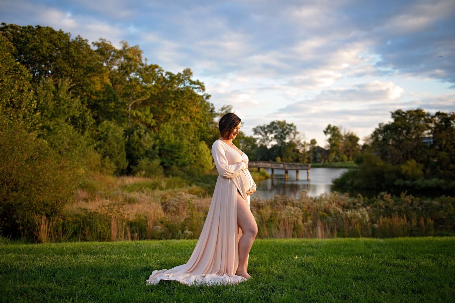 A maternity portrait in a pink flowing dress against a scenic lake background at sunset.