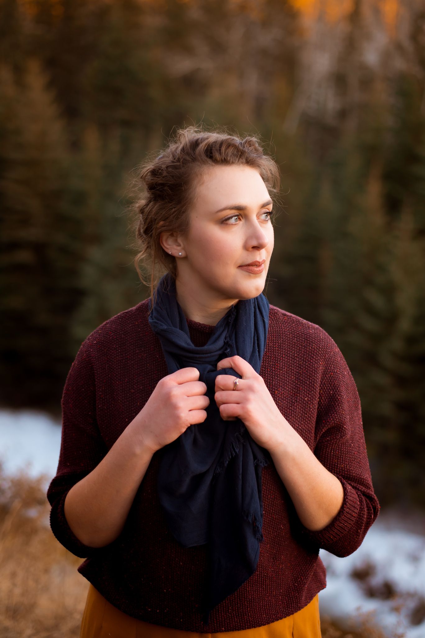 Person wearing a burgundy sweater and navy scarf gazes to the side during golden hour in a snowy winter setting.