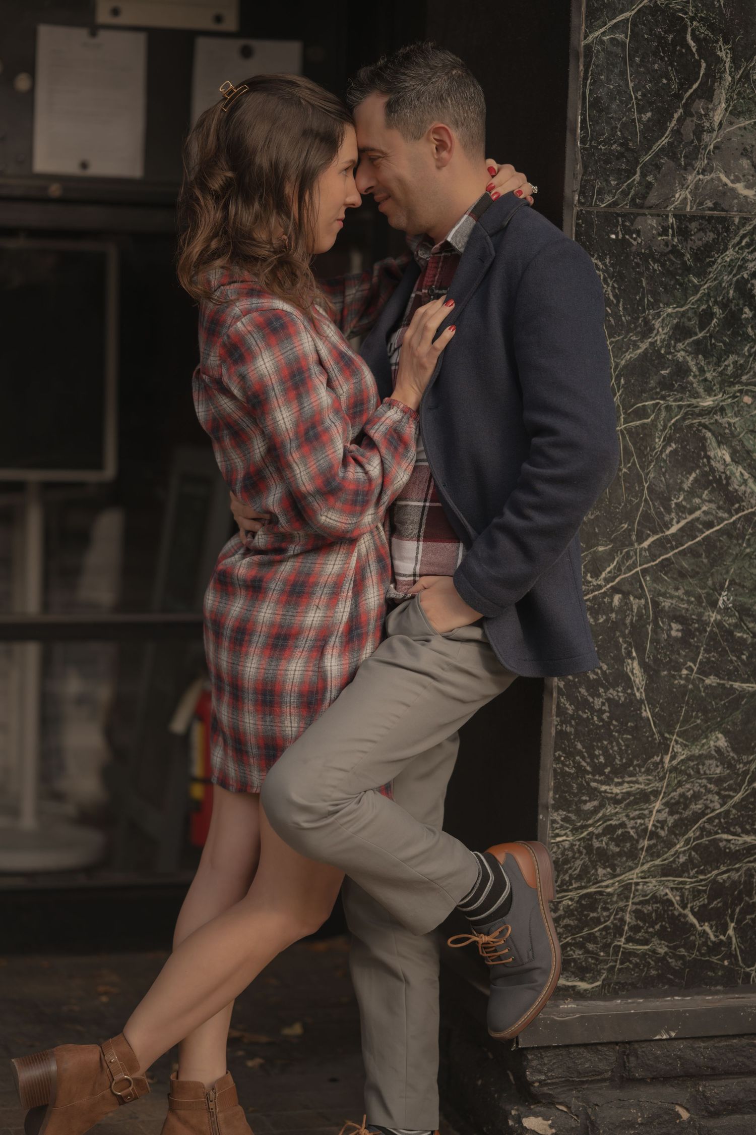 A romantic couple share an intimate moment against a wall, she in a plaid shirt dress and he in a navy blazer.