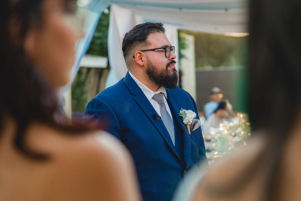 Bearded groom in a blue suit with glasses watching his bride approach during an outdoor wedding ceremony.