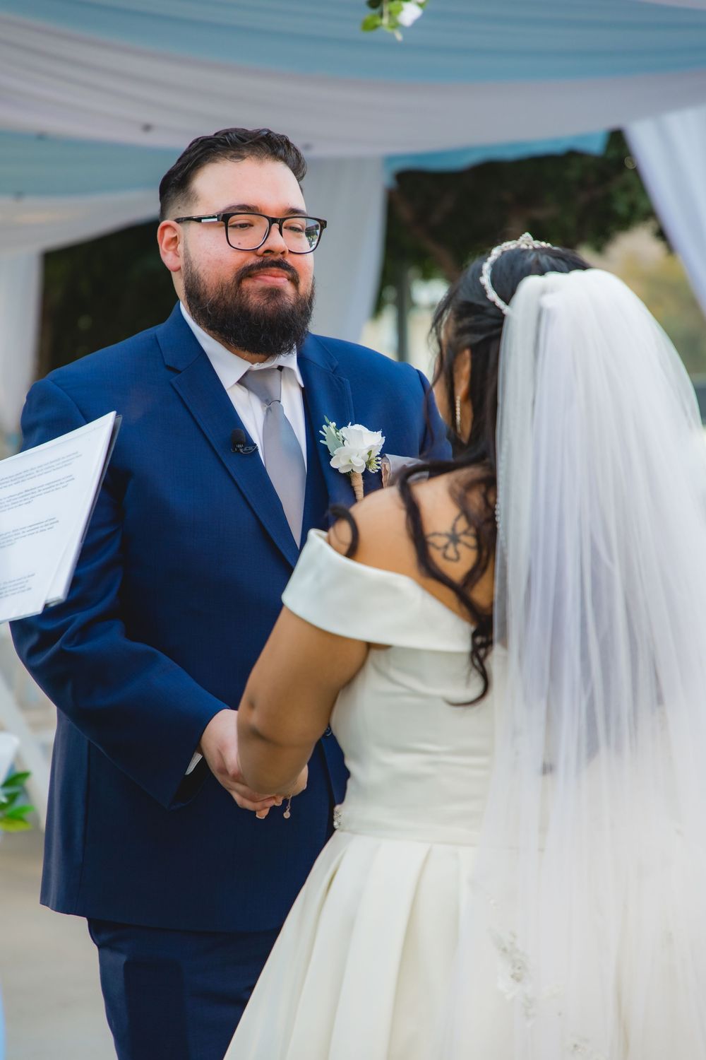 Groom in blue suit reading vows while holding hands with bride in white off-shoulder gown at altar.