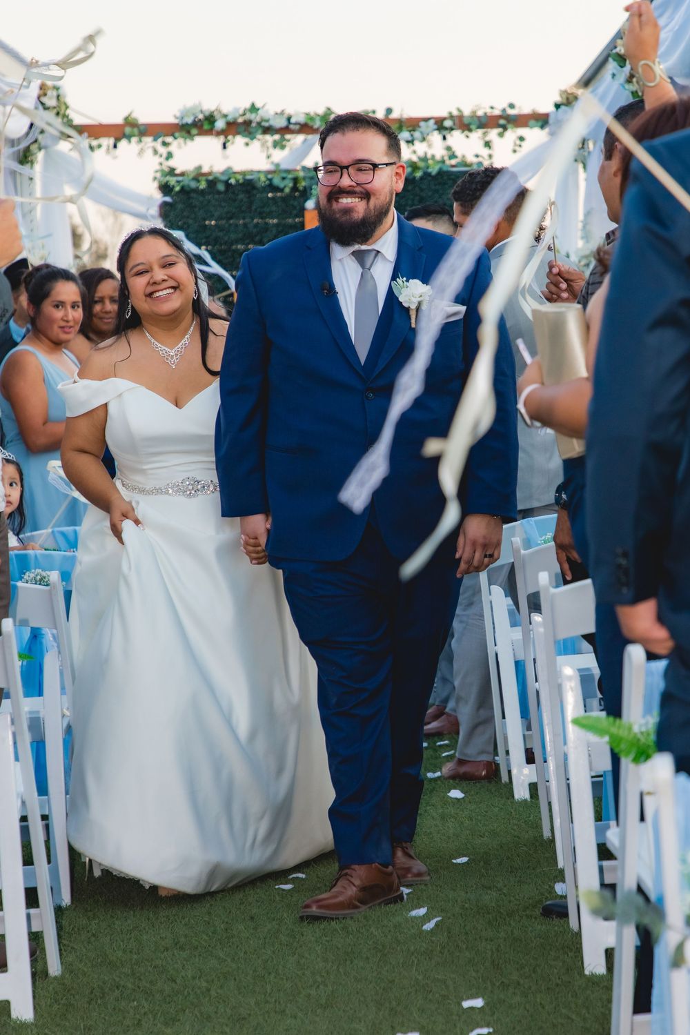 Joyful newlyweds walk down the aisle as guests wave ribbons at their outdoor wedding ceremony.