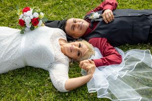 Newlywed couple lies on green grass holding hands with bridal bouquet beside them.