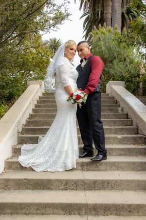 Bride in white lace gown and groom in burgundy shirt pose on stone staircase outdoors.