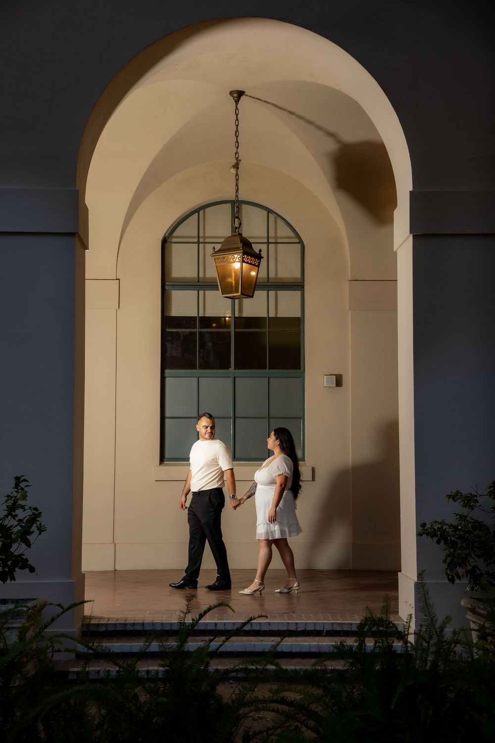 Couple holds hands while walking under an elegant arched portico with a vintage lantern at night.