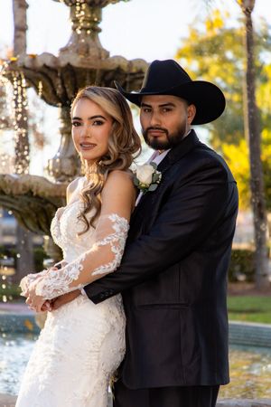 Bride and groom posing elegantly by a fountain, she in lace gown, he in black suit with cowboy hat.