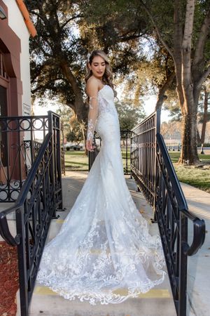 Bride in stunning lace mermaid wedding gown with long train standing on outdoor staircase surrounded by trees.