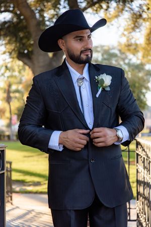 Dapper groom in black western suit with cowboy hat and boutonniere poses outdoors before wedding.