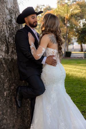 Groom in black cowboy hat leaning against tree embracing bride in lace wedding dress outdoors.