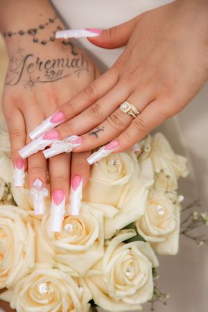 Close-up of bride's hands with pink and white acorn nails and diamond ring resting on white rose bouquet.